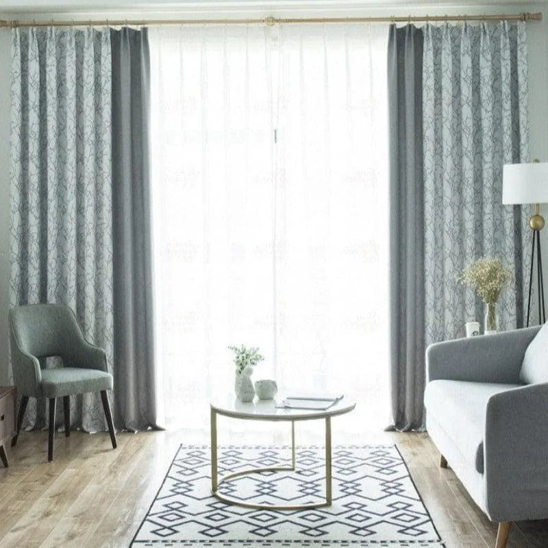 Salon moderne et élégant avec rideaux gris et imprimés géométriques, fauteuil confortable, canapé contemporain et table basse design sur un tapis à motifs. Ambiance lumineuse et déco minimaliste.