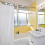 Salle de bain lumineuse avec murs carrelés jaunes et baignoire, équipée d'une vasque moderne. Rideau de douche blanc contrastant avec les tons chauds des murs. Fenêtre apportant une lumière naturelle abondante, créant une ambiance fraîche et accueillante. Design épuré et fonctionnel idéal pour optimiser l'espace et embellir votre intérieur.