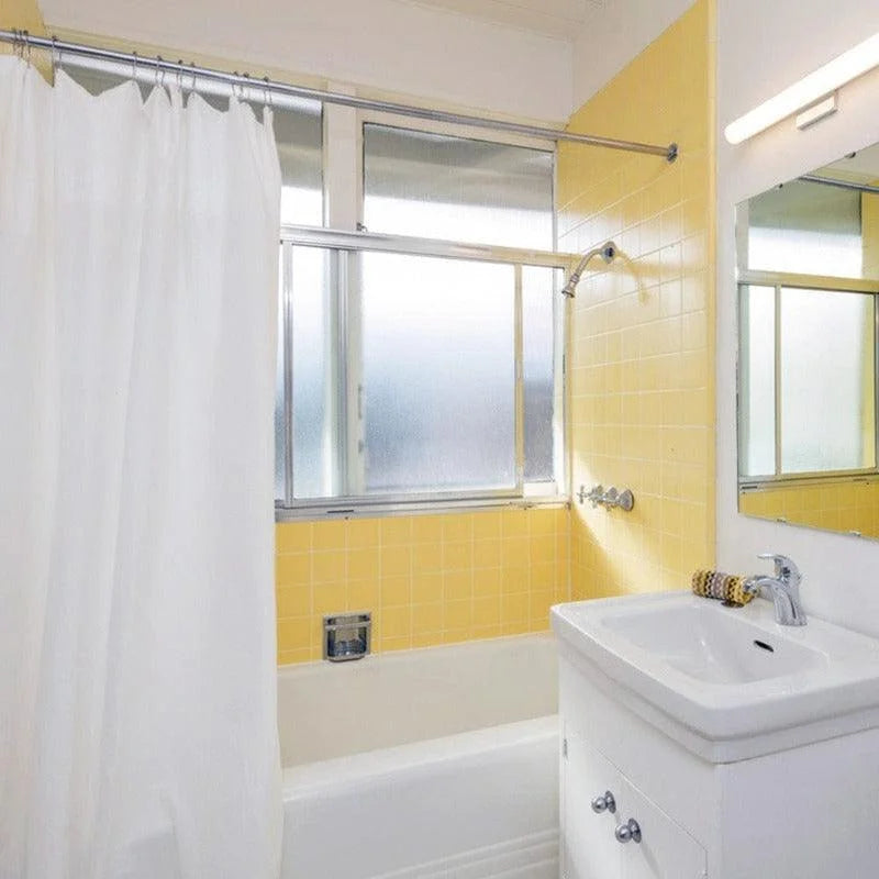 Salle de bain lumineuse avec murs carrelés jaunes et baignoire, équipée d'une vasque moderne. Rideau de douche blanc contrastant avec les tons chauds des murs. Fenêtre apportant une lumière naturelle abondante, créant une ambiance fraîche et accueillante. Design épuré et fonctionnel idéal pour optimiser l'espace et embellir votre intérieur.