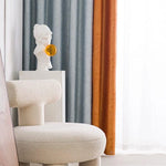 Fauteuil design moderne et élégant dans un intérieur chic, avec rideaux colorés en arrière-plan apportant une touche chaleureuse et artistique. Statue décorative sur un piédestal ajoutant une aura artistique et contemporaine au décor. Idéal pour un salon raffiné ou un espace de travail créatif.