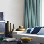 Salon moderne et élégant avec rideaux turquoise, canapé confortable orné de coussins bleus. L'espace est agrémenté de vases décoratifs en métal et d'une table basse avec livres, offrant une ambiance chaleureuse et contemporaine. Décoration intérieure sophistiquée et harmonieuse.