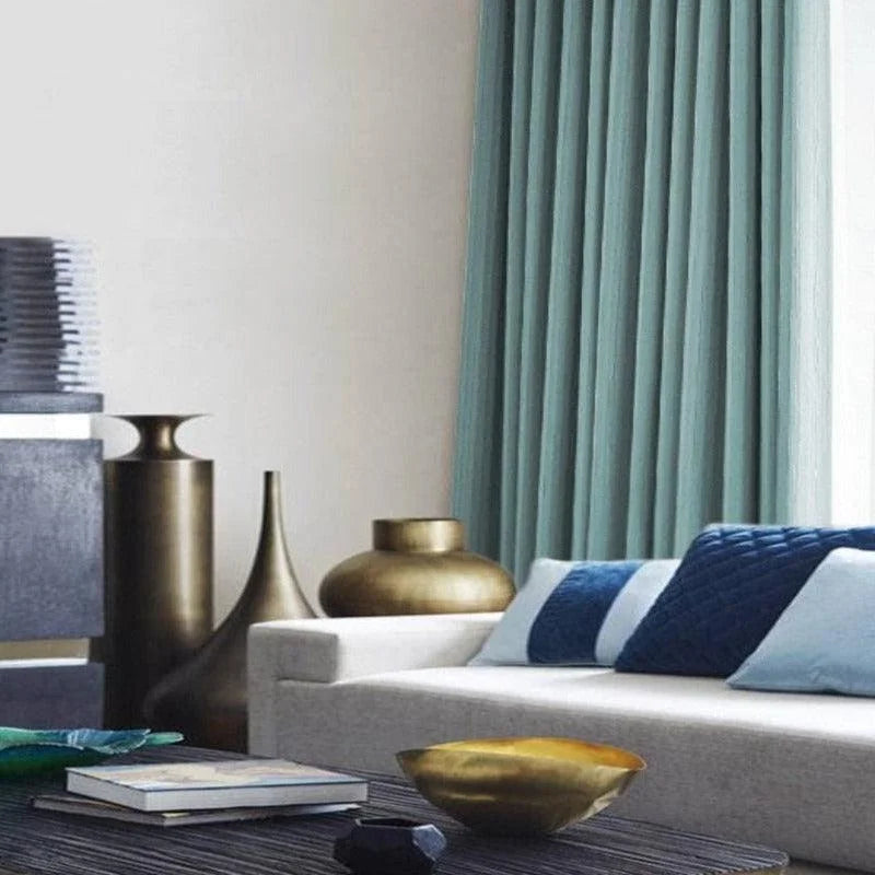Salon moderne et élégant avec rideaux turquoise, canapé confortable orné de coussins bleus. L'espace est agrémenté de vases décoratifs en métal et d'une table basse avec livres, offrant une ambiance chaleureuse et contemporaine. Décoration intérieure sophistiquée et harmonieuse.