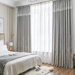 Chambre élégante avec lit double confortable, décor modernisé par de longues rideaux gris ornés de motifs subtils. Ambiance lumineuse grâce à une grande fenêtre laissant entrer la lumière naturelle. Décoration intérieure raffinée avec éléments dorés et texture douce au sol pour un espace accueillant et sophistiqué.