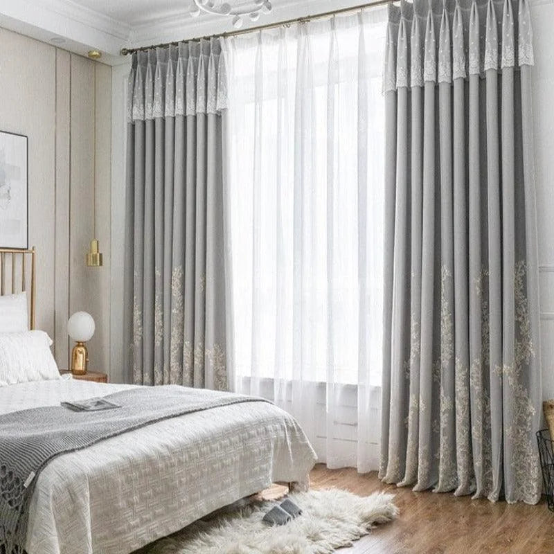 Chambre élégante avec lit double confortable, décor modernisé par de longues rideaux gris ornés de motifs subtils. Ambiance lumineuse grâce à une grande fenêtre laissant entrer la lumière naturelle. Décoration intérieure raffinée avec éléments dorés et texture douce au sol pour un espace accueillant et sophistiqué.