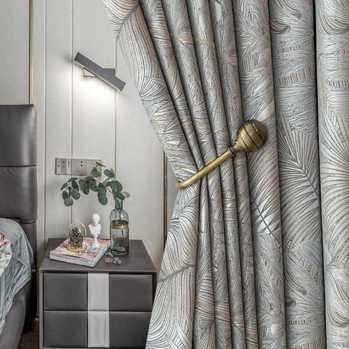 Rideaux modernes avec motif foliaire, ajoutant une touche élégante à la décoration intérieure d'une chambre contemporaine. L'agencement avec une embrasse dorée souligne le style raffiné tout en permettant de moduler la luminosité selon les envies. Parfaits pour apporter du cachet à votre espace de vie.