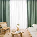 Salon moderne avec décor minimaliste. Rideaux verts et blancs en arrière-plan, meubles en bois clair, fauteuil et canapé confortables. Table basse avec vase et livres, ambiance lumineuse et apaisante idéale pour la détente et la lecture.