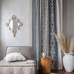 Salon élégant avec rideaux ornés de motifs géométriques, canapé moderne beige avec coussin duveteux, table d'appoint en bois, miroir mural design, et vase décoratif avec herbes séchées, créant une ambiance chic et apaisante pour votre intérieur.