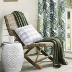 Fauteuil de terrasse confortable avec un plaid vert et un coussin à carreaux, placé près d'une table d'appoint blanche ajourée et d'un rideau à motifs tropicaux, idéal pour créer un coin détente extérieur élégant et accueillant.