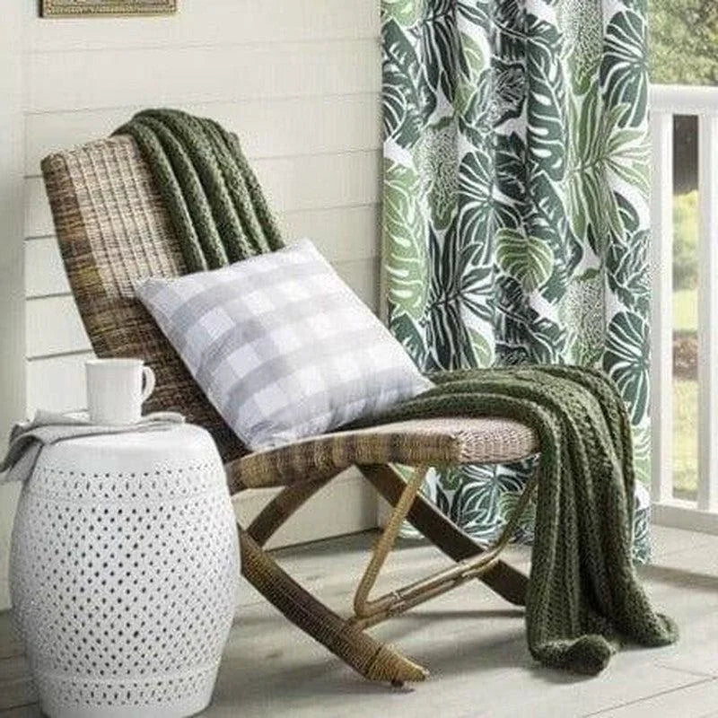 Fauteuil de terrasse confortable avec un plaid vert et un coussin à carreaux, placé près d'une table d'appoint blanche ajourée et d'un rideau à motifs tropicaux, idéal pour créer un coin détente extérieur élégant et accueillant.