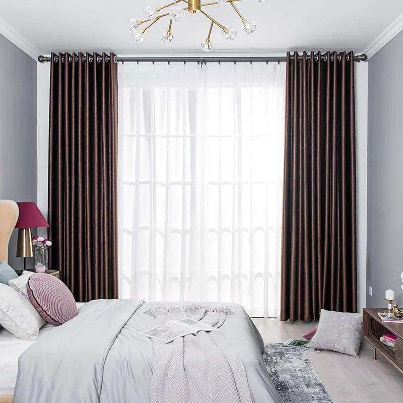 Chambre élégante avec rideaux longs et sombres encadrant une grande fenêtre, apportant une touche sophistiquée à la décoration intérieure. Le lit est joliment habillé avec des coussins et un couvre-lit assortis, offrant un espace accueillant et relaxant. Le mobilier moderne et les accessoires raffinés complètent le look luxueux et chaleureux. Parfait pour créer une ambiance reposante et stylistique dans toute chambre à coucher.