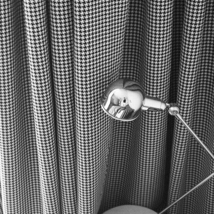 Lampe design en métal chromé posée devant un rideau à motif pied-de-poule noir et blanc. Accessoire élégant pour un intérieur moderne, apportant une touche de sophistication et un éclairage directionnel optimal. Parfait pour un bureau ou un coin lecture.