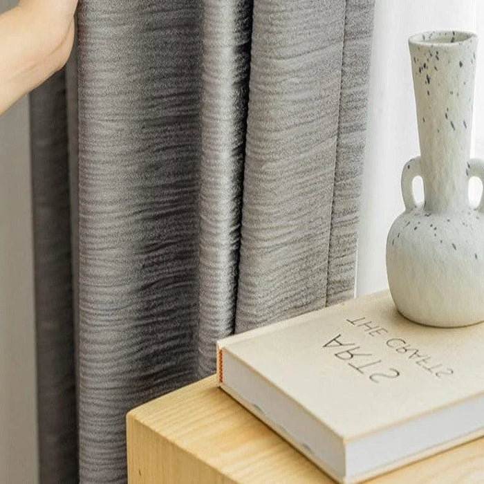 Rideau gris élégant dans un intérieur moderne, posé près d'une table en bois avec un vase design et un livre décoratif posé dessus. Idéal pour ajouter une touche contemporaine à votre salon ou chambre. Transformez votre espace avec ce rideau chic et fonctionnel.
