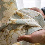 Image de rideau élégant à motifs floraux dans des tons de vert et or, tenu par une main, idéal pour l'intérieur et ajoutant une touche de sophistication à votre décoration.