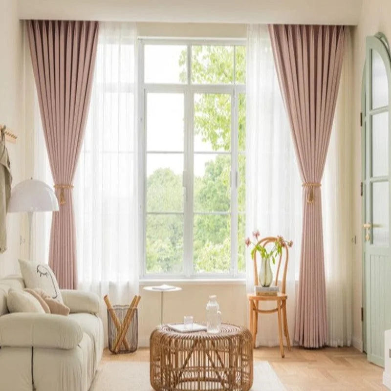 Salon lumineux avec de grandes fenêtres habillées de rideaux élégants couleur vieux rose. Ambiance chaleureuse et moderne grâce à la décoration intérieure harmonieuse comprenant un canapé douillet, une table basse en rotin, et une chaise décorative ajoutant une touche de charme. Idéal pour profiter de moments de détente en famille.
