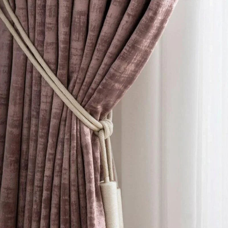 Rideau élégant de couleur rose poudre, replié avec un embrasse de cordon beige. Ce rideau habille une fenêtre en ajoutant une touche de sophistication et de style chaleureux à l'intérieur de la pièce. Idéal pour créer une ambiance cosy et chic dans le salon ou la chambre.