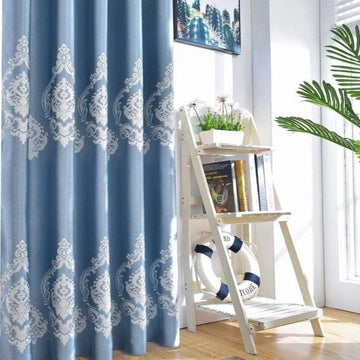 Rideau bleu élégant avec motifs décoratifs blancs, idéal pour apporter une touche sophistiquée et apaisante à votre salon. Parfait pour filtrer la lumière et créer une ambiance chaleureuse. Accessoirisé d'une étagère blanche avec des livres et des éléments décoratifs pour compléter l'ensemble du décor intérieur.