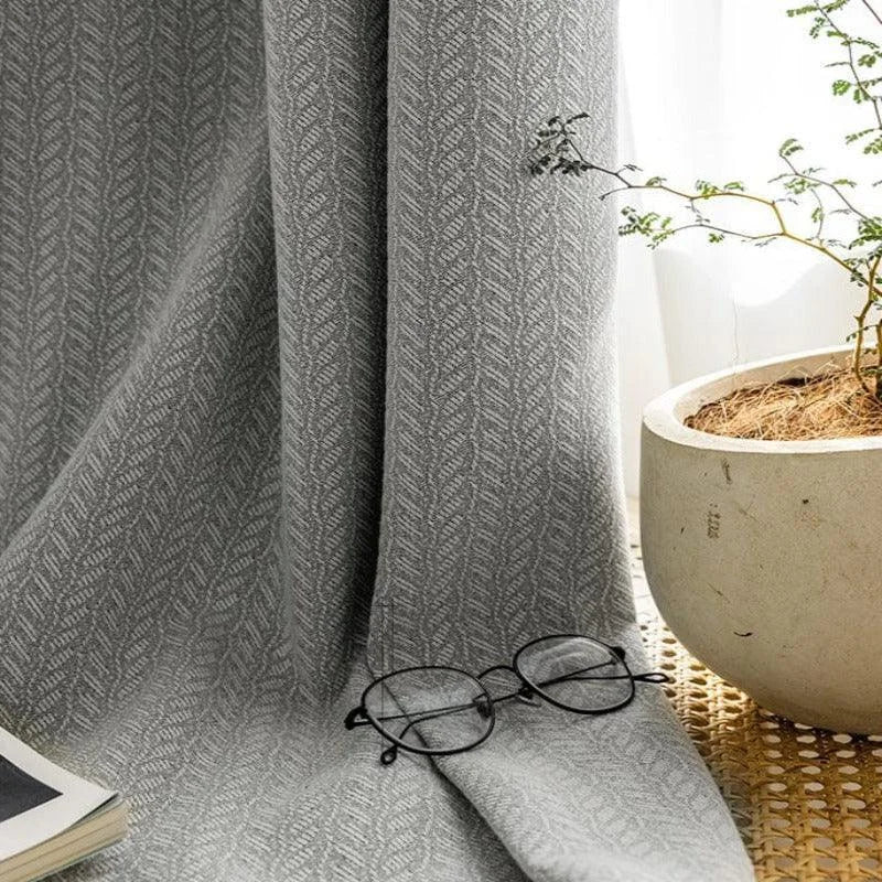 Rideau élégant de couleur grise avec motif géométrique, drapé devant une fenêtre lumineuse. Accessoirisé avec des lunettes noires et un livre fermé pour une ambiance de lecture relaxante. Une plante d'intérieur dans un pot blanc ajoute une touche naturelle à la scène. Idéal pour un salon moderne au style épuré.