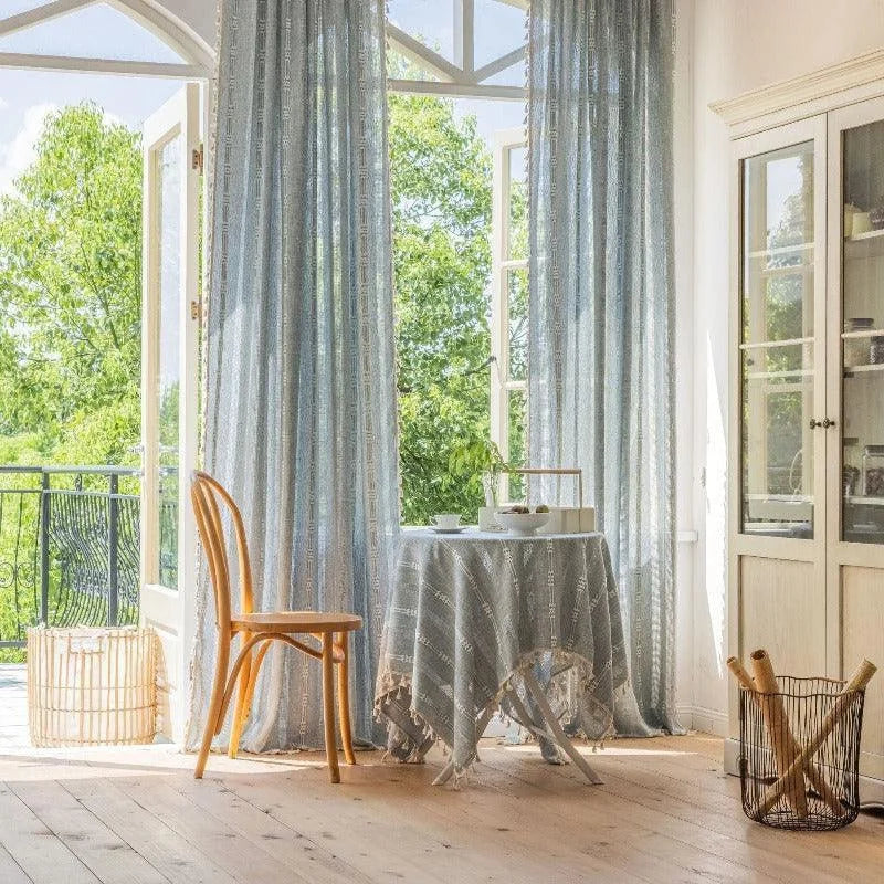 Salle à manger lumineuse avec table ronde décorée d'une nappe élégante à motifs devant de grandes fenêtres ouvertes donnant sur un jardin luxuriant. Ambiance chaleureuse avec une chaise en bois clair et de longs rideaux élégants qui filtrent la lumière naturelle. Éléments de décoration intérieure dans un espace invitant et paisible.