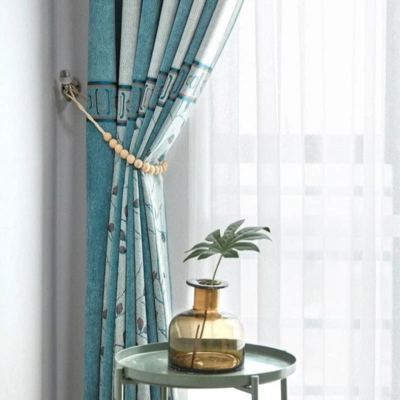 Rideau bleu élégant avec motifs décoratifs, attaché par un embrasse perlé. Devant, une table d'appoint moderne verte, ornée d'un vase en verre ambré contenant une feuille élégante, baignée de lumière naturelle. Idéal pour un salon raffiné, ajoutant une touche de style et de modernité à tout intérieur.