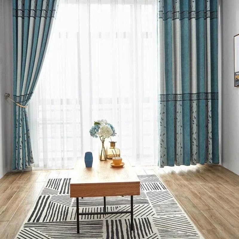 Rideaux élégants bleu et blanc ornant une baie vitrée, créant une ambiance lumineuse et moderne dans le salon. Table basse en bois stylée sur un tapis graphique, décorée avec des accessoires contemporains. Idéal pour un intérieur raffiné et accueillant.