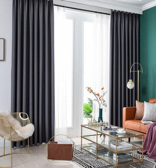 Salle de séjour moderne avec une ambiance élégante, mettant en valeur de longs rideaux de couleur noire qui ajoutent une touche de sophistication. Le canapé orange vif contraste harmonieusement avec le mur vert, créant un espace convivial et accueillant. Une table basse en verre avec des finitions dorées trône au centre de la pièce, entourée d'un fauteuil design et d'éléments décoratifs raffinés. L'éclairage naturel inonde la pièce grâce aux grandes fenêtres, accentuant le