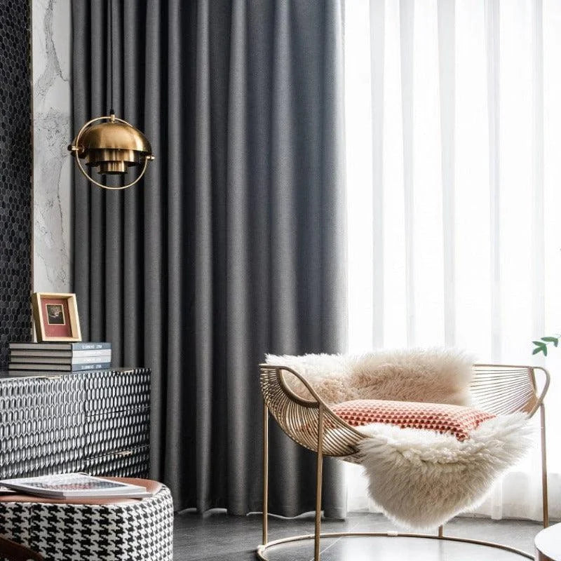 Fauteuil élégant au design moderne, idéal pour salon contemporain. Décoration intérieure avec rideaux sombres contrastés par des voilages blancs, lampe suspendue dorée, et éléments décoratifs tendance. Atmosphère chic et confortable, adaptée pour un coin lecture ou relaxation.