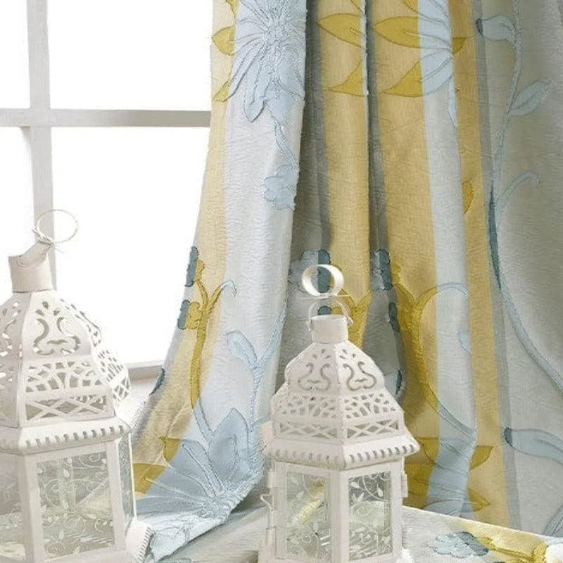 Rideau floral aux teintes douces de bleu et jaune, ajoutant une touche élégante et lumineuse. Deux lanternes blanches au design raffiné posées devant, parfaites pour une décoration intérieure accueillante et chaleureuse. Fenêtre en arrière-plan laissant filtrer une douce lumière naturelle. Idéal pour transformer l'ambiance d'un salon ou d'une chambre.