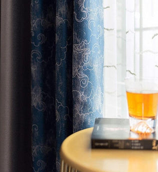Rideaux élégants aux motifs nuageux accentués de bleu, apportant une touche artistique à votre intérieur. Un verre ambré posé sur une table en bois à côté d'un livre, prête un air de détente et d'harmonie à cet espace lumineux. Parfait pour ajouter du style à votre salon.