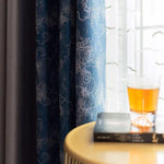 Rideaux élégants aux motifs nuageux accentués de bleu, apportant une touche artistique à votre intérieur. Un verre ambré posé sur une table en bois à côté d'un livre, prête un air de détente et d'harmonie à cet espace lumineux. Parfait pour ajouter du style à votre salon.