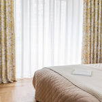 Chambre élégante avec literie en harmonie et rideaux à motifs floraux, lumière naturelle filtrant à travers les voilages blancs, parquet en bois au sol. Idéal pour une ambiance cosy et sereine.