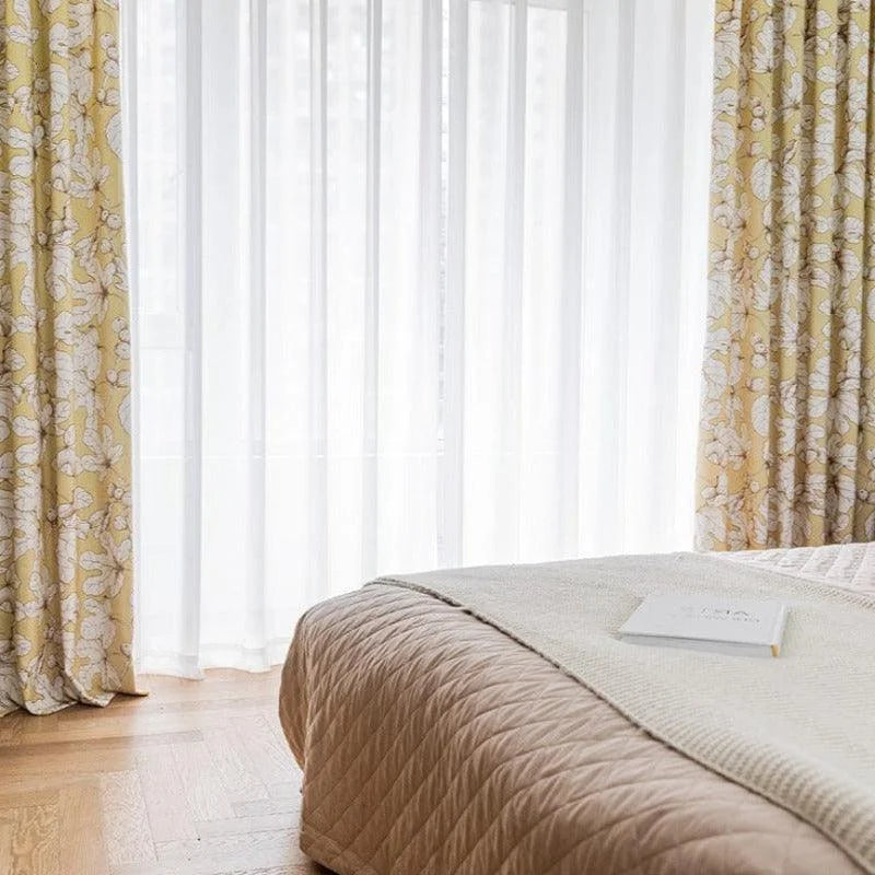 Chambre élégante avec literie en harmonie et rideaux à motifs floraux, lumière naturelle filtrant à travers les voilages blancs, parquet en bois au sol. Idéal pour une ambiance cosy et sereine.