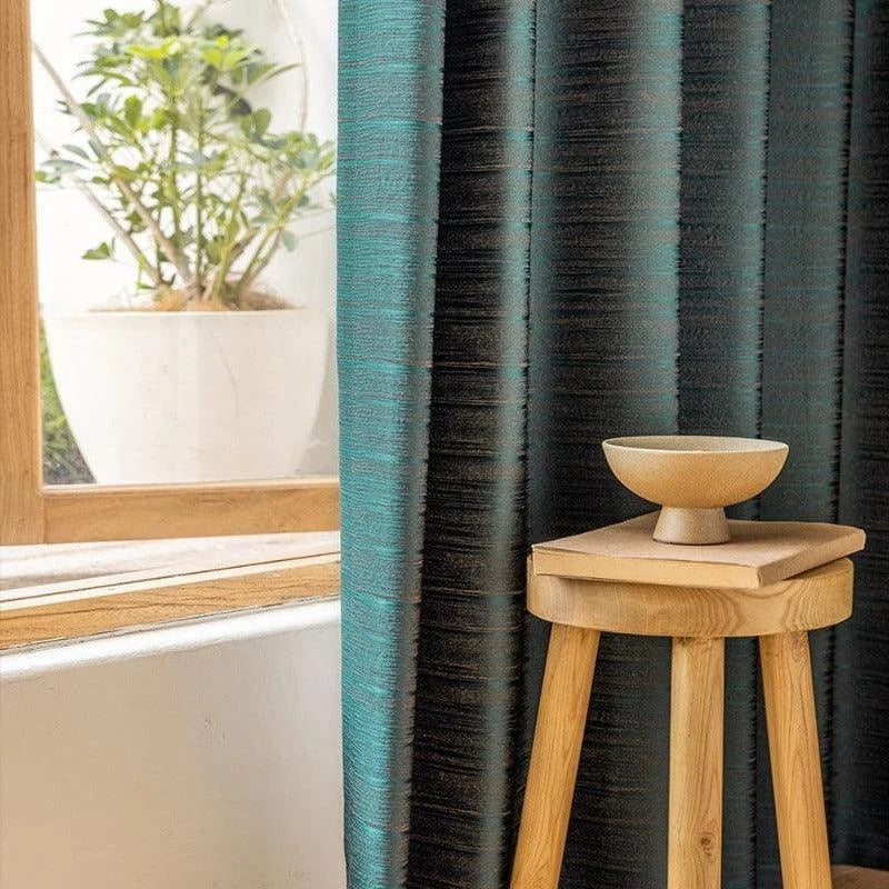 Rideau moderne bleu et noir dans un intérieur élégant avec lumière naturelle, accompagné d'un tabouret en bois naturel surmonté d'un bol décoratif. Fenêtre en bois et plante d'intérieur en arrière-plan créant une atmosphère chaleureuse et apaisante. Parfait pour un salon contemporain.