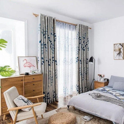 Chambre moderne et lumineuse avec rideaux à motifs floraux bleus, offrant une atmosphère apaisante. Décor scandinave avec une chaise en bois, commode en bois clair et lampe de chevet design. Ensemble harmonieux dans une pièce élégante et contemporaine.