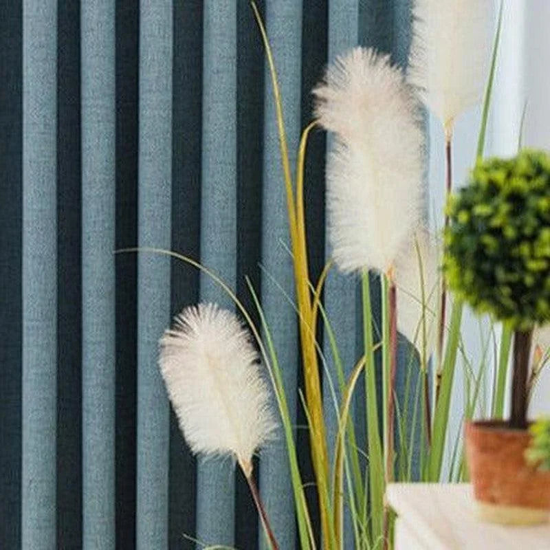 Rideau plissé bleu foncé derrière des herbes décoratives blanches et vertes, à côté d'une plante en pot sur une table blanche, créant une ambiance moderne et apaisante pour votre intérieur.