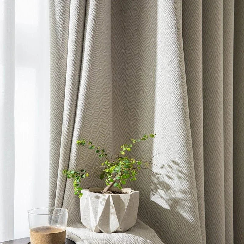 Rideau beige texturé avec motif chevrons, apportant une ambiance élégante et contemporaine à votre intérieur. Parfait pour créer une atmosphère chaleureuse et lumineuse. Accompagné d'une plante en pot et d'un verre, ce décor est idéal pour mettre en valeur un salon ou une chambre moderne.