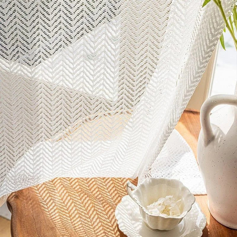 Voilage élégant avec motif chevrons, laissant passer la lumière naturelle pour une ambiance chaleureuse. Idéal pour le salon ou la chambre. Sur la table, une tasse avec soucoupe blanche ajoute une touche d'élégance.