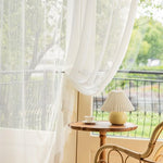 Voilage blanc donnant sur un balcon avec vue verdoyante, créant une ambiance lumineuse et apaisante dans un salon. Petite table en bois avec une lampe élégante et une tasse, ajoutant une touche de confort et de style à la décoration intérieure.