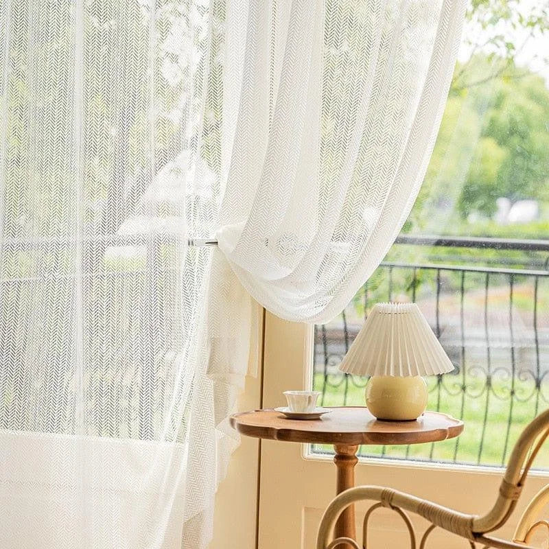 Voilage blanc donnant sur un balcon avec vue verdoyante, créant une ambiance lumineuse et apaisante dans un salon. Petite table en bois avec une lampe élégante et une tasse, ajoutant une touche de confort et de style à la décoration intérieure.
