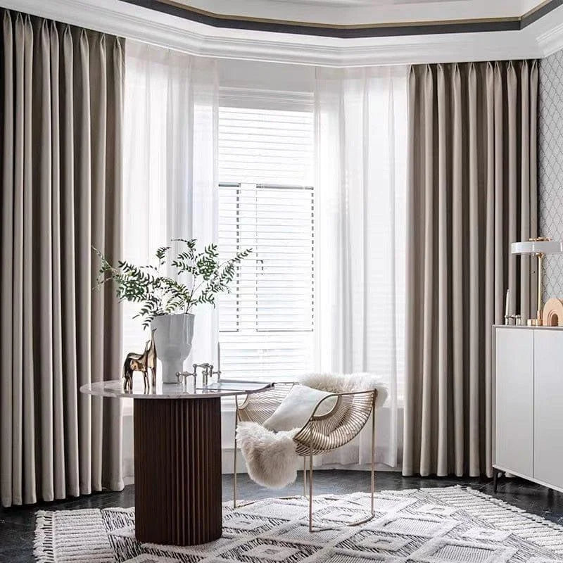 Salle élégante avec de grands rideaux beige créant une ambiance apaisante. Décor moderne avec une table ronde, une chaise design dotée d'un coussin doux, et un vase avec des feuillages verts. Tapis aux motifs géométriques ajoutant du style à l'espace lumineux.