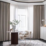 Salle élégante avec de grands rideaux beige créant une ambiance apaisante. Décor moderne avec une table ronde, une chaise design dotée d'un coussin doux, et un vase avec des feuillages verts. Tapis aux motifs géométriques ajoutant du style à l'espace lumineux.