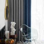 Chaise design transparente placée dans un salon moderne avec rideaux élégants gris et bleus. Un miroir rond doré orne le mur aux côtés d'un buste artistique sur une table en bois. La lumière naturelle illumine la pièce à travers les rideaux translucides, offrant un espace lumineux et accueillant. Aménagement sophistiqué et art déco pour un intérieur contemporain.