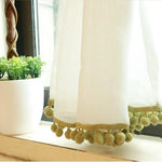 Voilage élégant avec pompons verts pour décoration intérieure, idéal pour ajouter une touche raffinée à votre salon ou chambre. Rideau léger suspendu près d'une fenêtre, accompagné d'une plante en pot sur une table, créant une ambiance chaleureuse et apaisante. Accessoire de maison moderne parfait pour filtrer doucement la lumière naturelle.