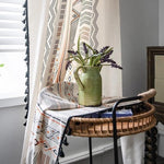 Rideaux à motifs géométriques colorés ajoutent une touche bohème au salon. Table en osier avec un vase vert et fleurs, éclairée par la lumière naturelle, créant une ambiance chaleureuse. Décoration intérieure élégante et contemporaine. Idéale pour embellir un espace avec style et modernité.