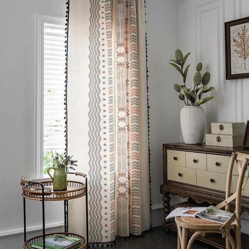 Rideau à motifs géométriques bohémiens, suspendu dans un salon lumineux. La scène inclut une table d'appoint en rotin avec une petite plante décorative verte, un tiroir vintage, et une chaise en bois naturel. L'ensemble crée une ambiance chaleureuse et élégante, idéale pour un intérieur moderne et stylé.