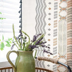 Rideau moderne aux motifs géométriques dans des tons noirs et orangés, apportant une touche élégante à votre intérieur. Associé à un pichet vert contenant un bouquet de fleurs, cet ensemble dégage une atmosphère chaleureuse et accueillante dans la pièce. Idéal pour un salon ou une chambre, ce rideau est un choix parfait pour personnaliser votre décoration intérieure avec style.