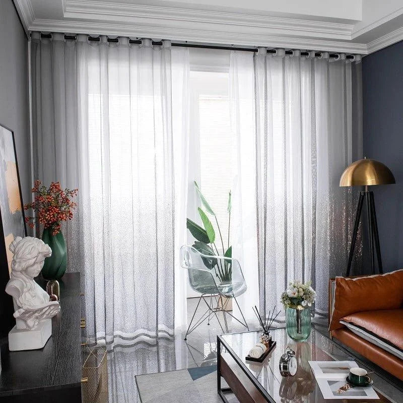 Salon moderne avec rideaux transparents, grande baie vitrée, canapé en cuir brun, lampe dorée sur pied, table basse en verre décorée et plante verte, créant une ambiance lumineuse et élégante. Idéal pour un intérieur design mettant en valeur la lumière naturelle et le style contemporain.