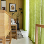 Salon moderne avec rideaux verts et mobilier en bois clair, créant une ambiance naturelle et accueillante. Plantes décoratives, lumière naturelle abondante, et décoration murale artistique ajoutent une touche d'élégance à la pièce. Idéal pour un intérieur contemporain et chaleureux.