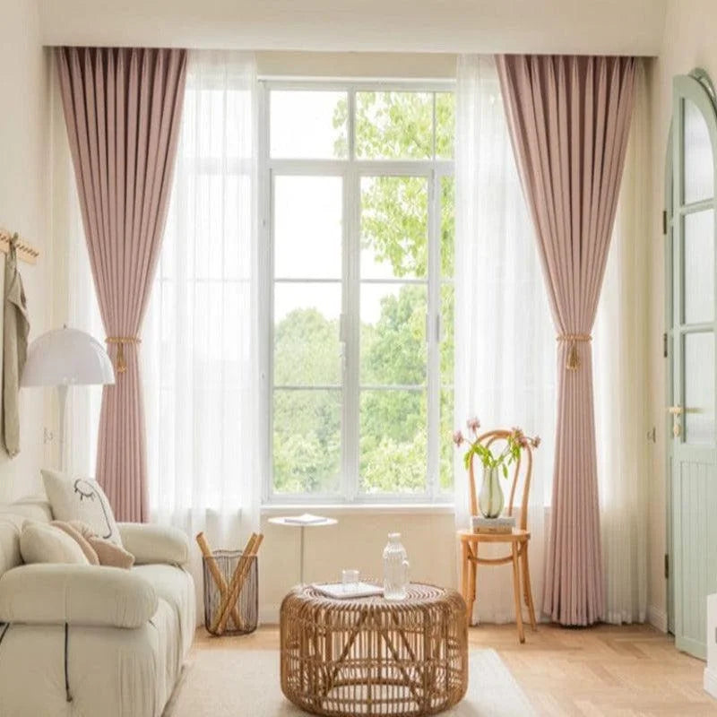 Salon moderne avec rideaux élégants roses encadrant une grande fenêtre laissant entrer la lumière naturelle. Décoration intérieure épurée avec table basse en rotin, canapé confortable et chaises design. Idéal pour aménager un espace convivial et lumineux dans votre maison.
