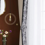 Élégant espace de vie avec rideaux à motif zèbre et éclairage design suspendu. Miroir doré accroché au mur chocolat, vase avec fleurs fraîches ajoutant une touche de couleur. Ambiance moderne et chic pour un intérieur raffiné.