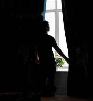 Homme devant une fenêtre ouverte, ajustant un rideau pour laisser entrer la lumière naturelle dans la pièce, avec une plante d'intérieur sur le rebord de la fenêtre, créant une ambiance de tranquillité et de bien-être à domicile.